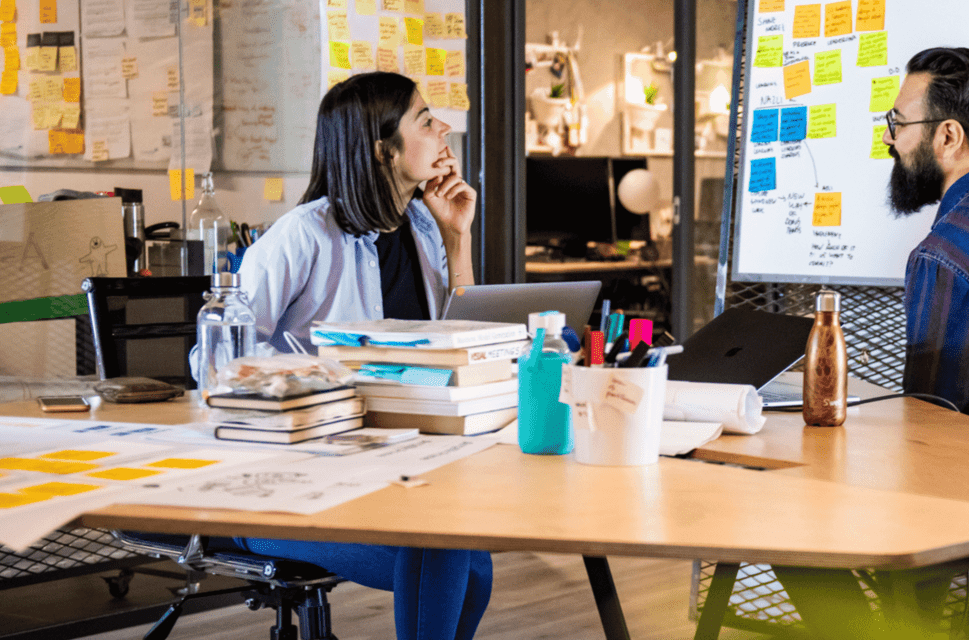 A woman and a man sit at a table in a creative hub. They are chatting and the walls of the room are covered in brightly coloured sticky notes.