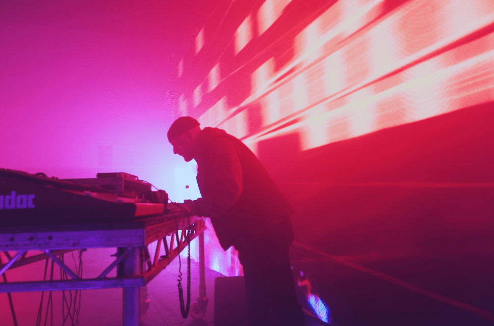 A DJ stands at a set of decks, lit from behind against a purple/pink and orange brightly lit backdrop. He leans forwards.