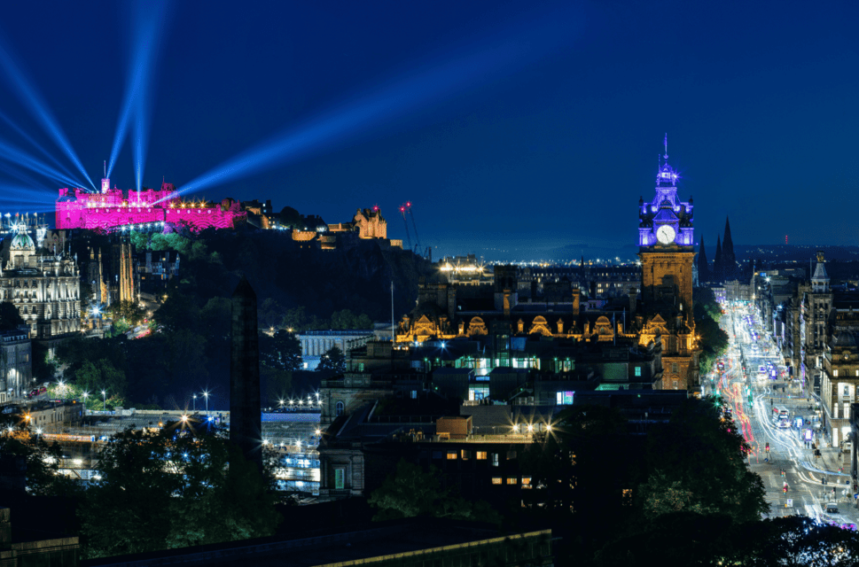 The Edinburgh skyline at night, with searchlights coming out of the castle on a hilltop, which is illuminated in pink light. A busy street is lit up to the right of the image.