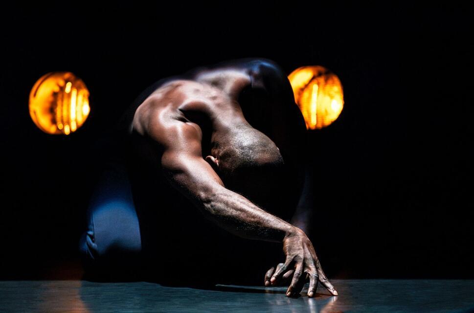 Dancer Enowate kneels on the stage floor in front of car headlights