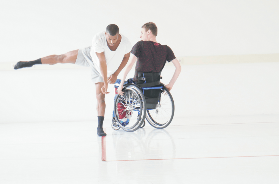 Two male performers stand next to each other in a white space. The man on the left stands in the foreground, bending forwards at the waist with his right leg outstretched. The performer behind and to his right uses a wheelchair and sits with his back to the camera.