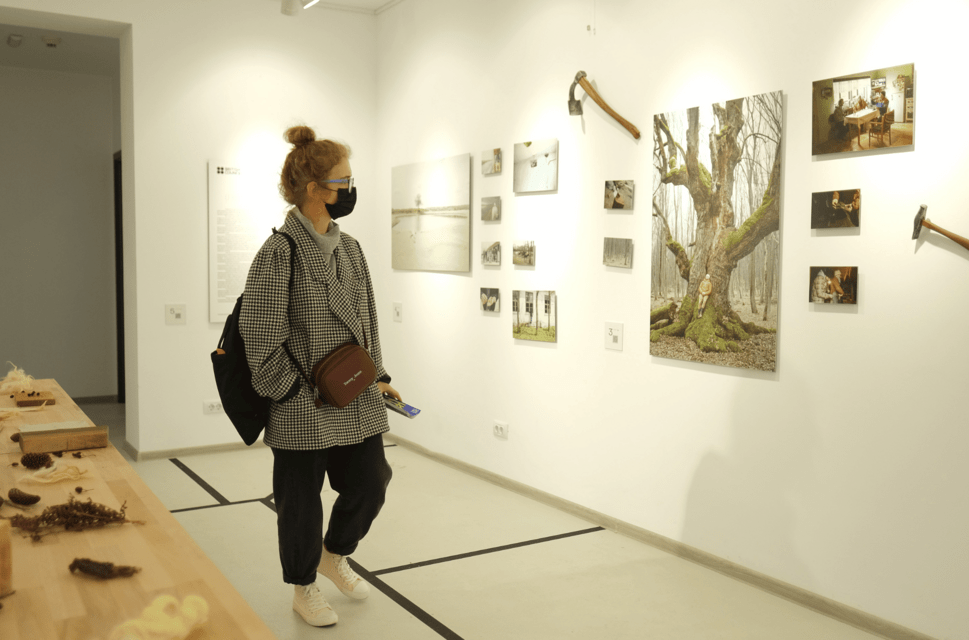 A woman in a face mask in a gallery space, looking at a wall-mounted display of tools and a painting of a tree.