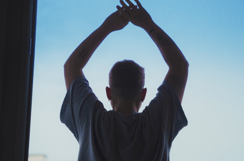 A young man stands in an open doorway, facing away from the viewer, silhouetted against a pale blue sky with his arms held up above his head, and his hands joined together.