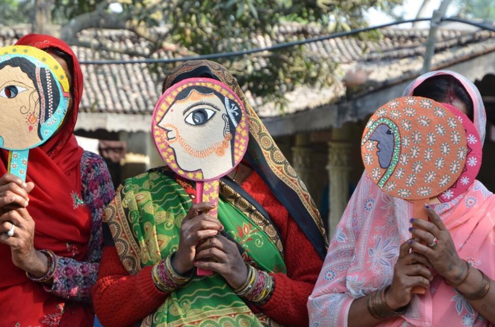 Three women in saris stand in a row with their faces obscured by hand-held masks on sticks with the faces of women painted on them.