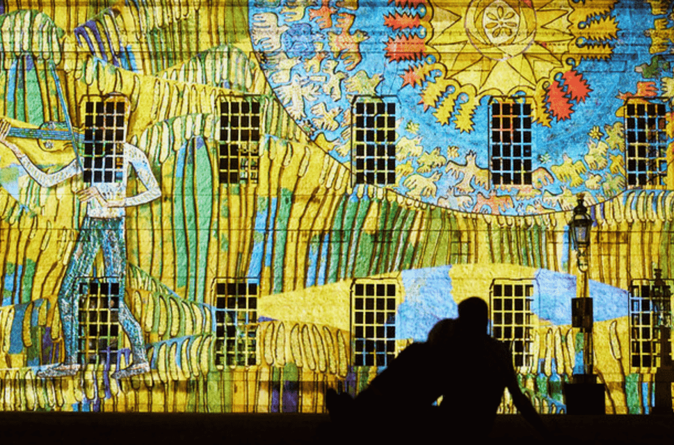 A projection of vibrant blues and yellows in an abstract pattern against the frontage of Buckingham Palace. A seated couple is silhouetted in the foreground, looking on.