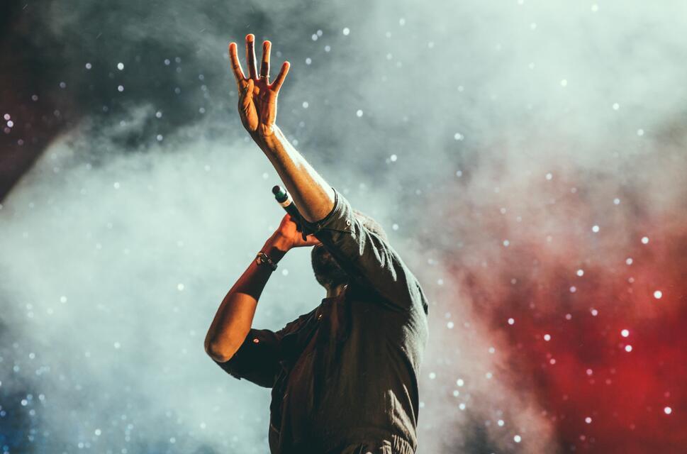 Man on stage with bright lights and white smoke, holding a microphone in one hand and raising four fingers into the air with the other.