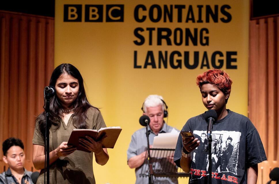 Two poets of Asian decent stand infront of a yellow banner at the BBC Contains Strong Language festival to perform their poems to a live audience.