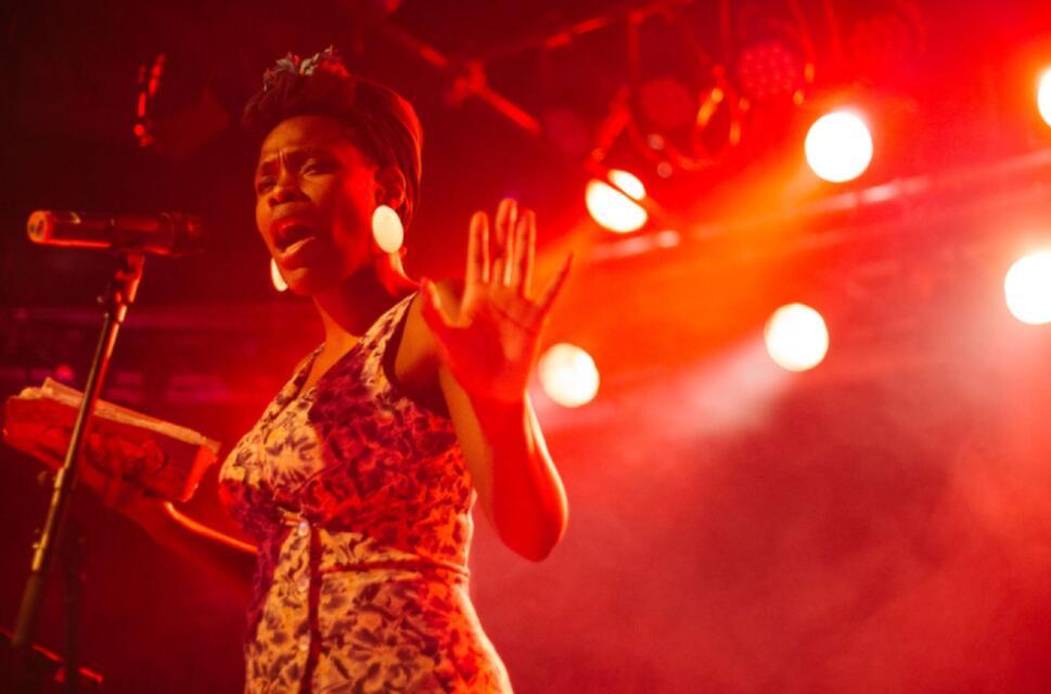 Vanessa Kisuule stands performing on stage infront of a microphone under red lights and smoke.