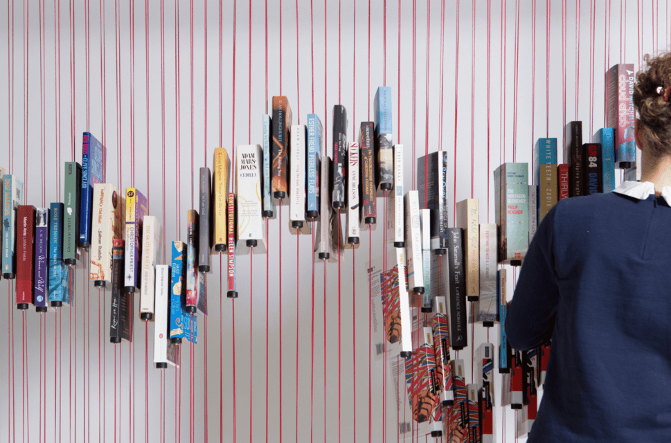 Paperback books suspended on vertical red strings at different heights, creating a zig-zag pattern. To the right of the image, a woman stands with her back to the camera, adding or removing books from the string ‘bookcase’