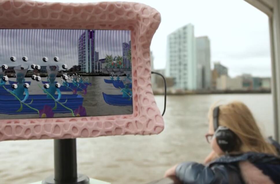 Digital screen on a boat showing animated fish over a city. On the boat, behind the sceen, there's a passenger wearing headphones looking out to the river.