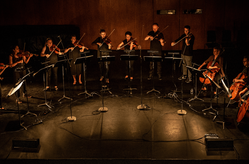A strings ensemble perform in a semi-circle on a dimly lit stage.