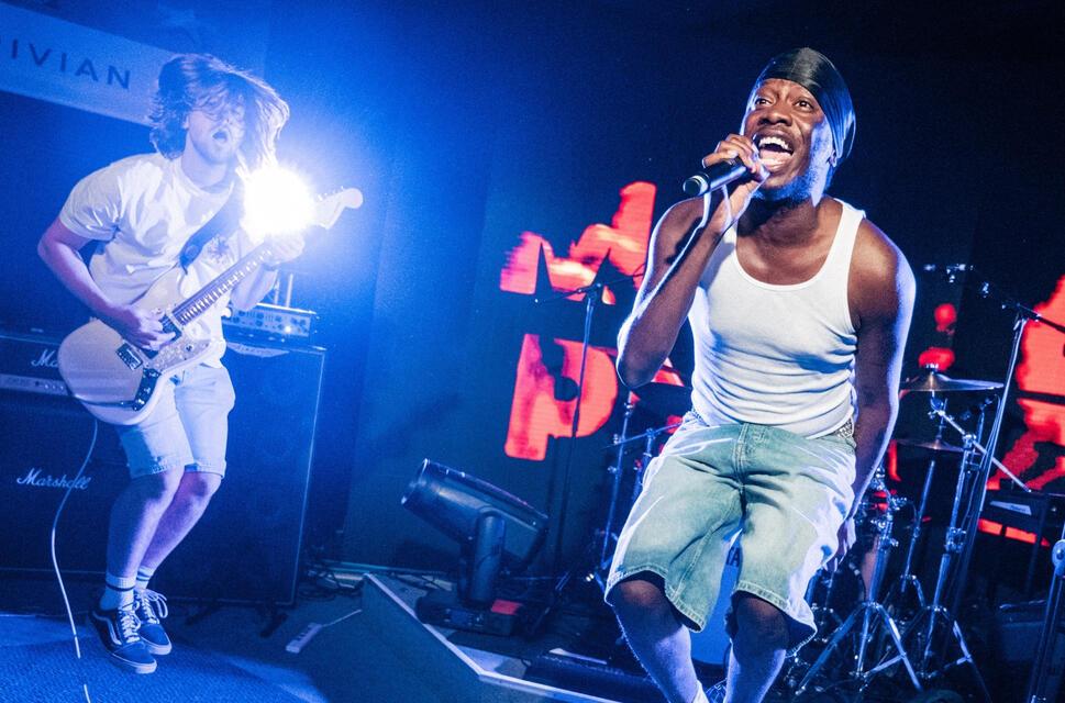 On stage, electric guitarist on left, jumping into the air; vocalist on right wearing white vest also jumping.