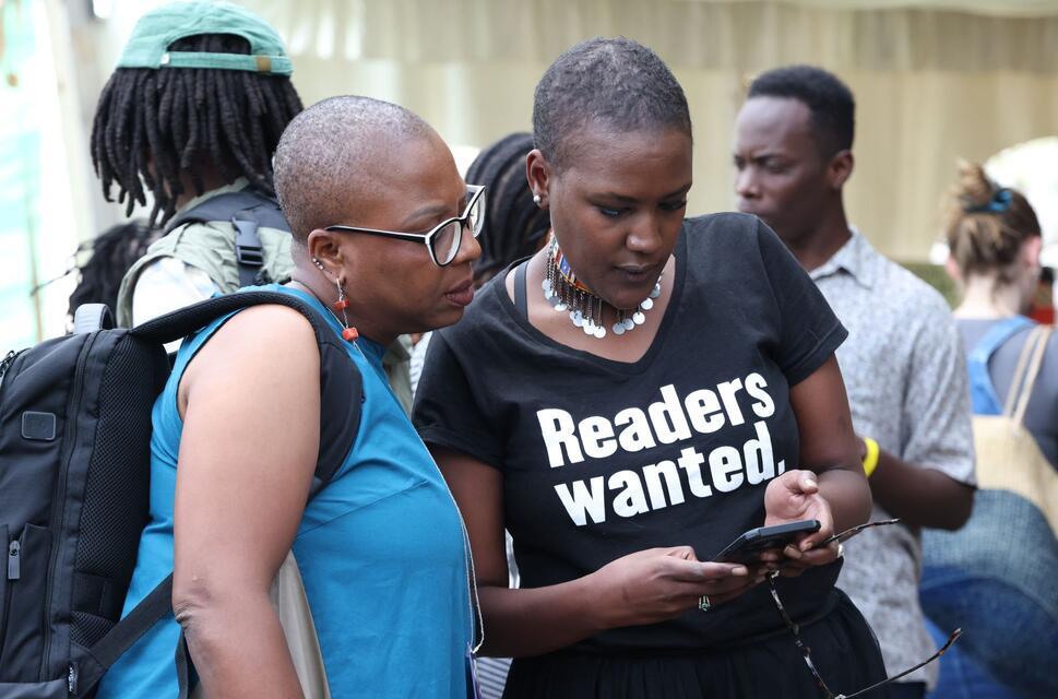 Festival goers, with one wearing a Readers Wanted t-shirt lean over a phone at the NBO Literature Festival 2024