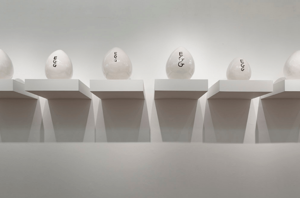 A row of small white shelves hold ceramic white eggs of different sizes, with the word 'EGG' painted in black vertically on each. The shelves and eggs are mounted against a white wall.
