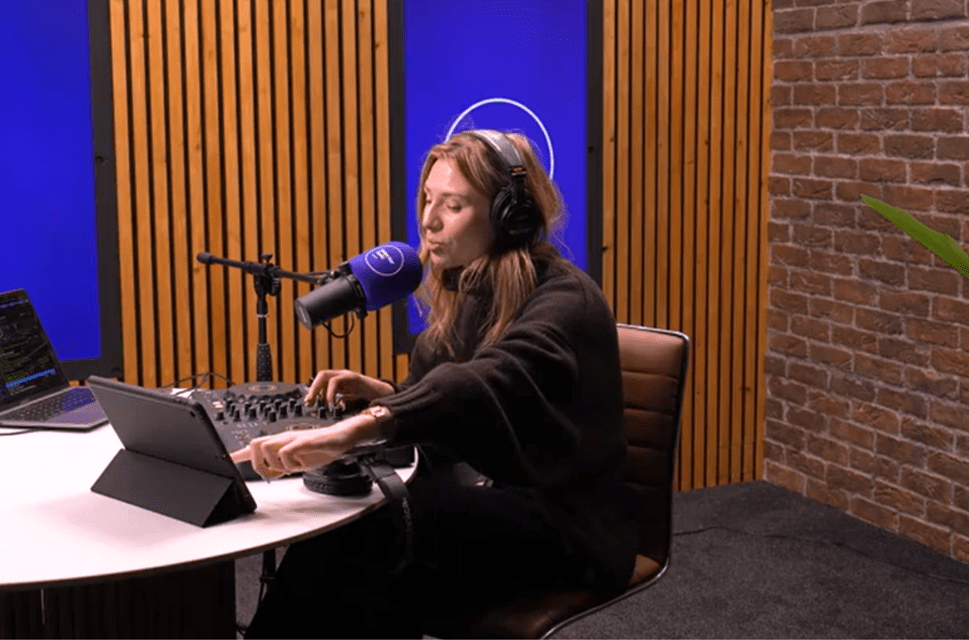 Lady in radio studio with headphones on, microphone in front of her, at a desk, two laptops next to her, blue screens behind her.