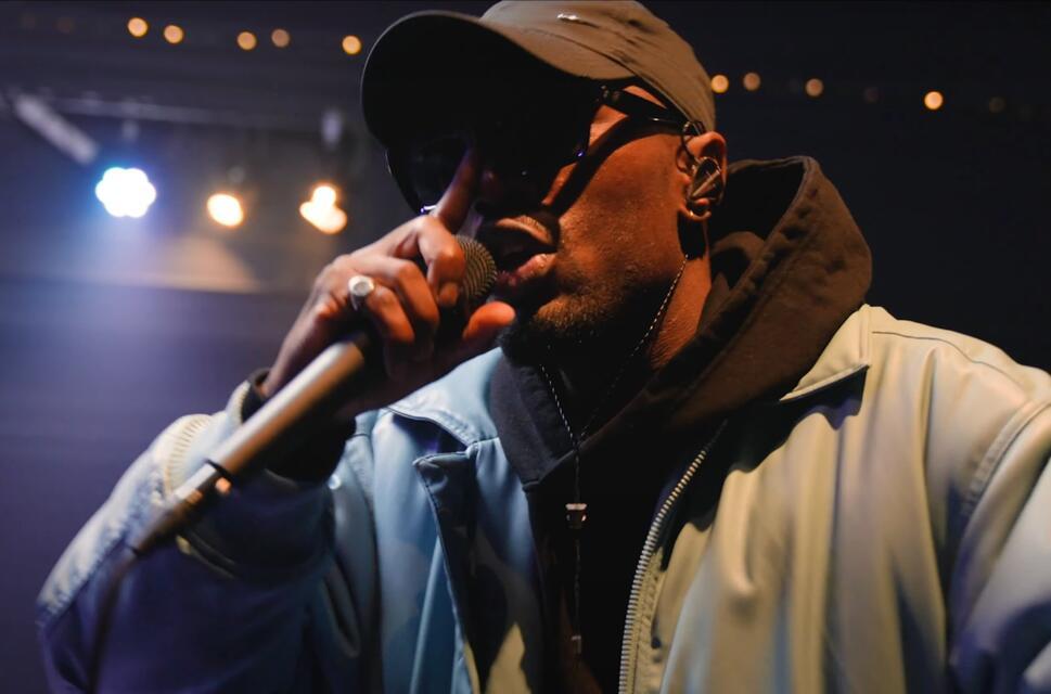 Close up of man, holding microphone to his mouth, wearing hooded top, cap and sunglasses. Bright indoor stage lights behind him.
