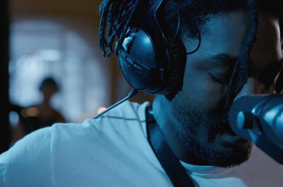 Man wearing headphones, looking down, behind microphone in dark studio setting.