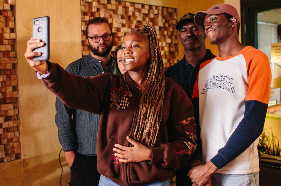 Five people in a recording studio, stood with the young lady at the front holding a camera to take a picture of them all.