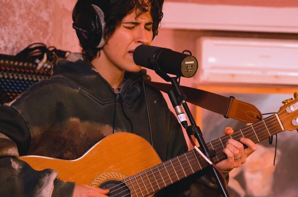 Someone in a studio, playing guitar, wearing headphones, stood behind microphone, eyes closed.