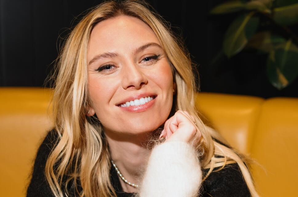 Close up of lady's face. She has blonde hair, sat on bright orange sofa, smiling.