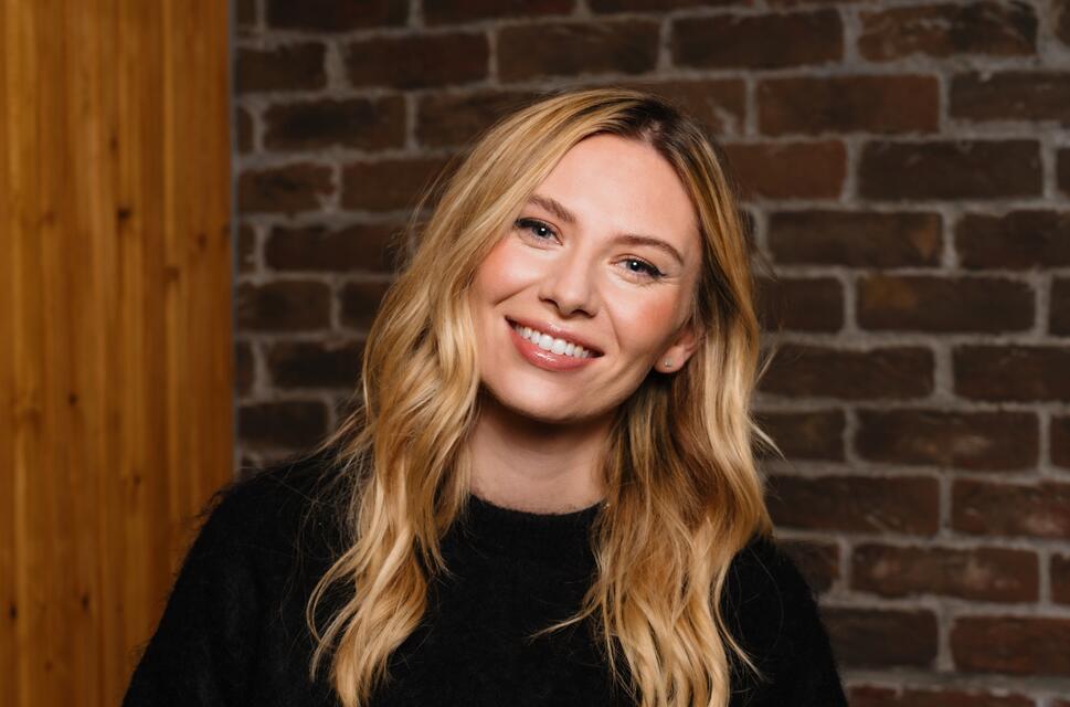 Lady with long blonde hair, smiling towards camera, in front of interior brick wall.