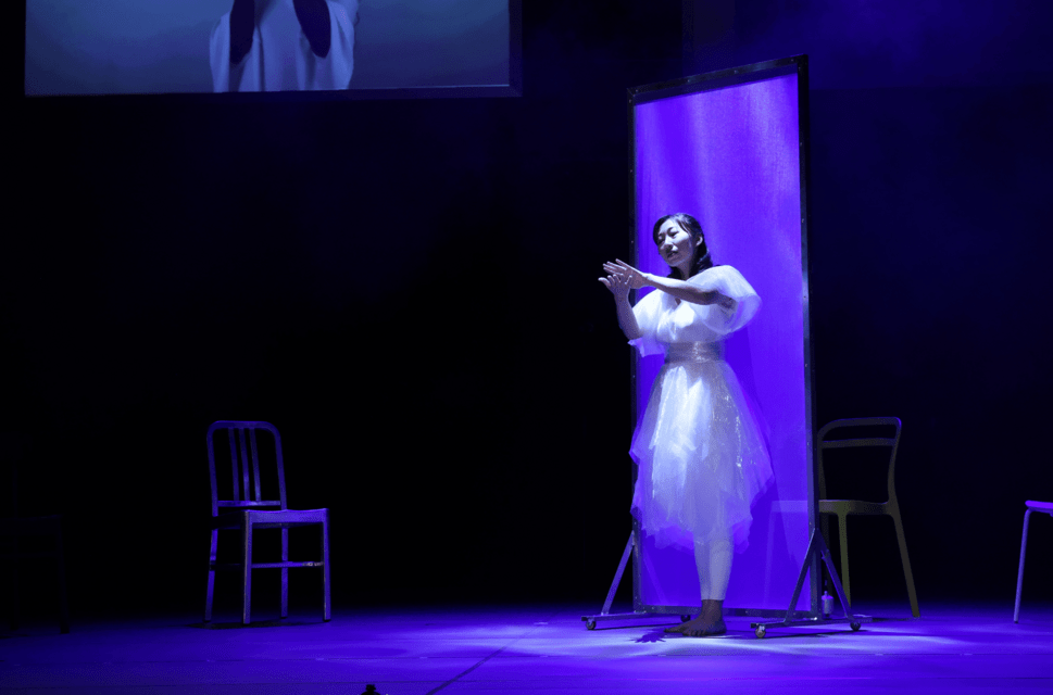 A female performer wearing a white dress stands on a stage dimly lit by blue-purple lighting. She is gesturing towards the audience with her left hand. She stands in front of a tall white screen and there are three chairs arranged around her. At the top left of the frame, the bottom of a screen or projection is just visible, which seems to show the bottom half of a figure in white with long dark hair.
