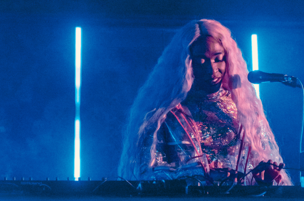 A Black woman, wearing a sequined outfit and with long pink hair, stands in front of a microphone and operates a sound desk. Behind her is a haze of dry ice and bright blue neon lights.