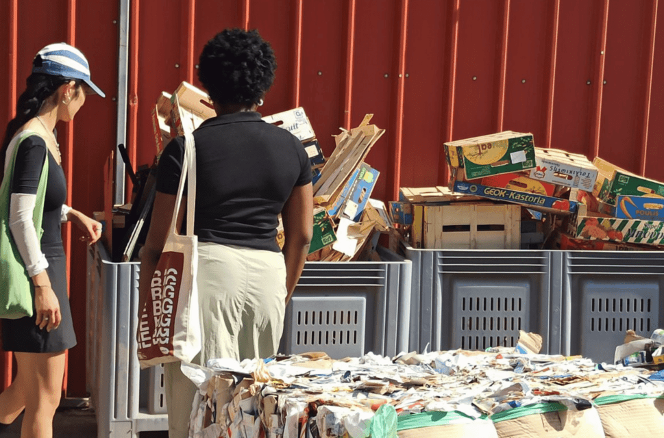 Two women stand with their backs to the camera, looking at a recycling unit which is overflowing with wooden crates and cardboard boxes. Cubes of compacted materials for recycling stand in the foreground, slightly out of focus