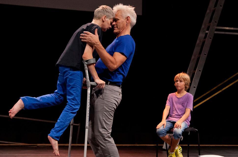 A performer using crutches leans in closely, forehead to forehead, with another adult performer in a tender moment on stage. A child sits nearby on a chair, watching. The scene is part of Claire Cunningham’s The Way You Look (at me) Tonight, performed at Southbank Centre’s Unlimited Festival.