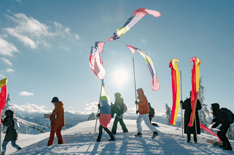 Eight figures walking across over snowy ground from right to left of the frame. They are wearing clothing for cold weather and are carrying colourful banners or flags on long poles.The sky is blue with a few small clouds and the sun is in the centre-background above the figures' heads.
