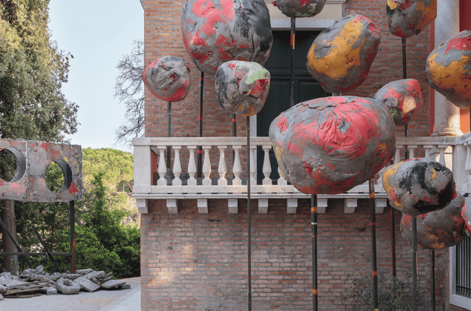 Large spherical papier mache sculptures of various sizes painted red, yellow and grey, mounted on sticks on the right/foreground of the image. Behind the sculptures the side of a redbrick building with a white balustrade can be seen. In the background on the left of the image stands another grey, red and yellow sculpture, which is shaped like a large placard mounted on two sticks, with two large circular holes cut into it. 