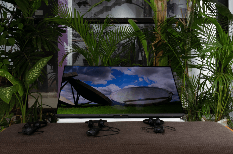 A plasma screen is set on top of a table, angled upwards. The screen shows images of satellite dishes against a bright blue sky with white clouds. In front of the screen there are headphone sets, and around the display are tropical leaved plants.