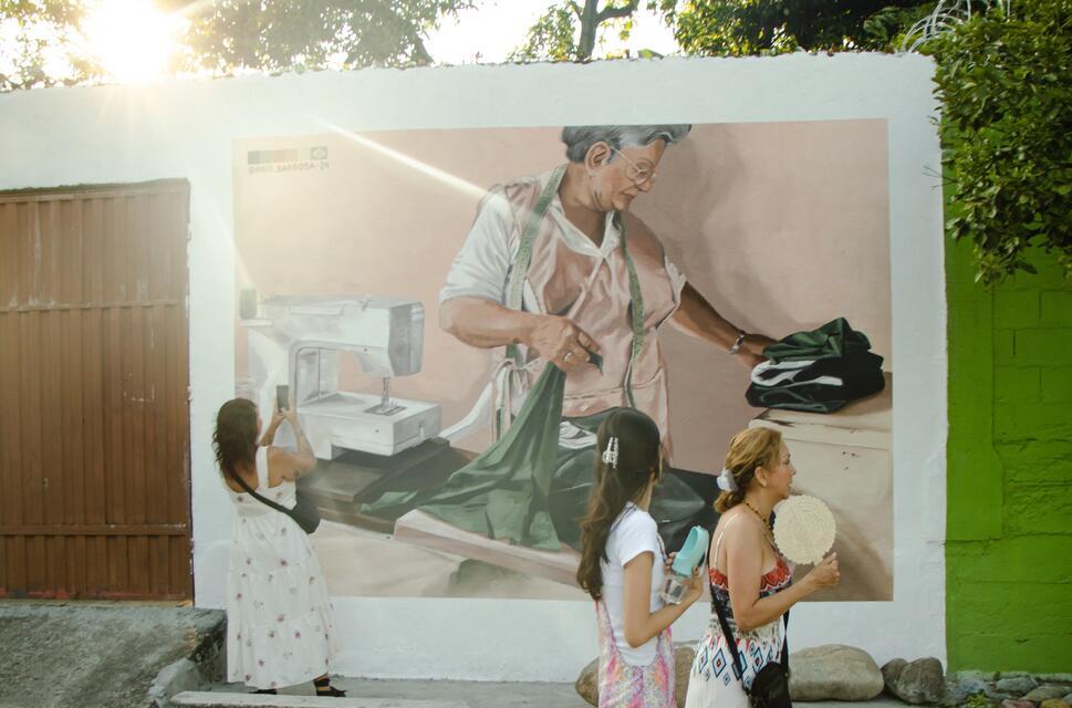 A painted mural shows a woman doing laundry on an outdoor wall. The sunsets behind the wall and passerbys look and take pictures.