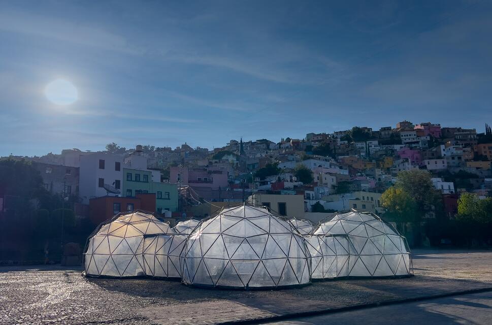 Transparent geodesic domes stand in a public square beneath a hillside cityscape under a bright blue sky.