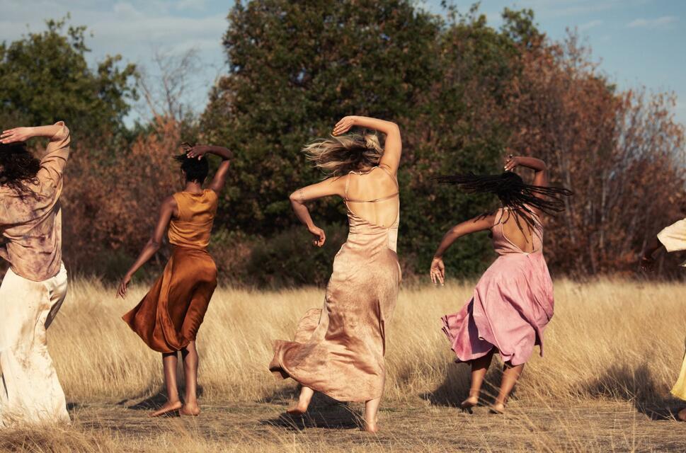 Five women in flowing dresses dance outside in nature